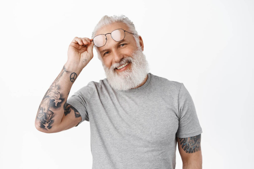 Homem grisalho tatuado sorrindo e levantando os óculos, exibindo dentes tratados com lentes de contato dental em Sumaré.