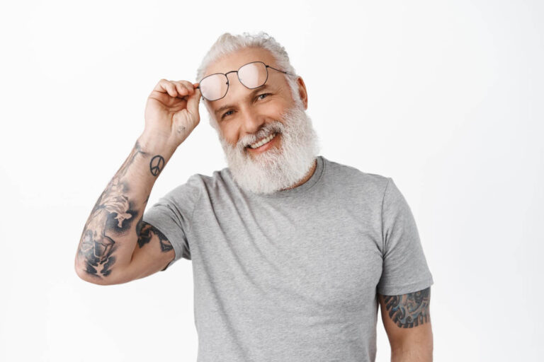 Homem grisalho tatuado sorrindo e levantando os óculos, exibindo dentes tratados com lentes de contato dental em Sumaré.