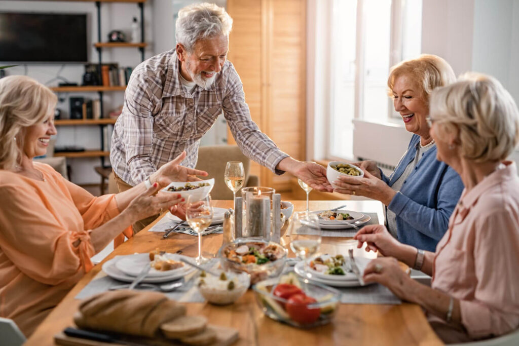 Grupo de amigos idosos sorrindo e compartilhando comida à mesa. Sinta o implante dentário como um dente natural em Sumaré.